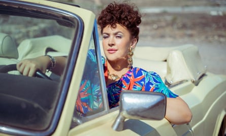 Una mujer vestida de forma llamativa conduce un descapotable de los años 80 bajo el sol.