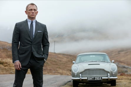 Daniel Craig como Jame Bond en Skyfall, 2012, parado al costado de una carretera con un auto deportivo detrás de él.
