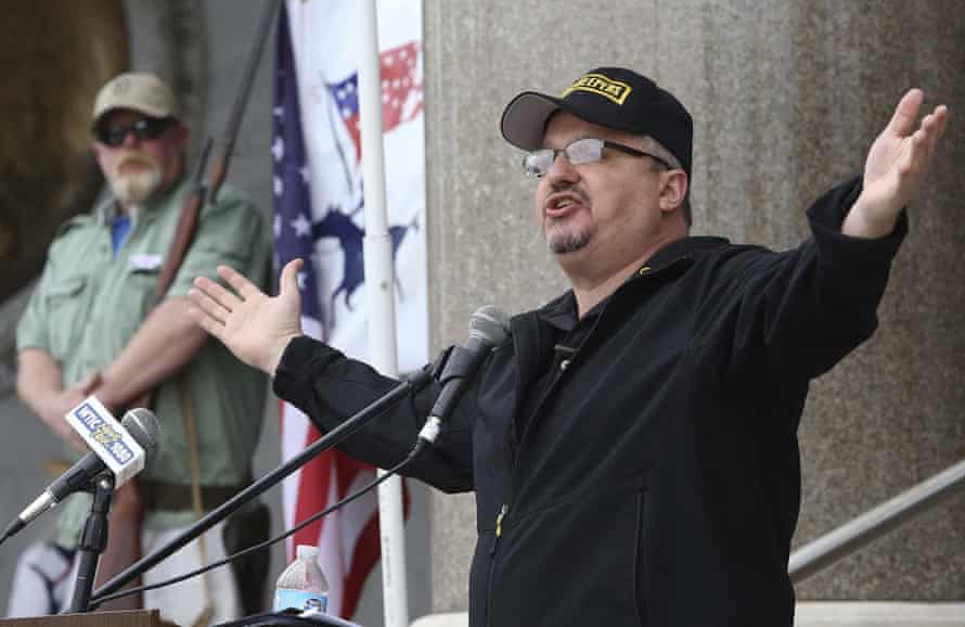 Un hombre vestido con una camisa negra y una gorra de béisbol se dirige a una multitud a través de un micrófono.